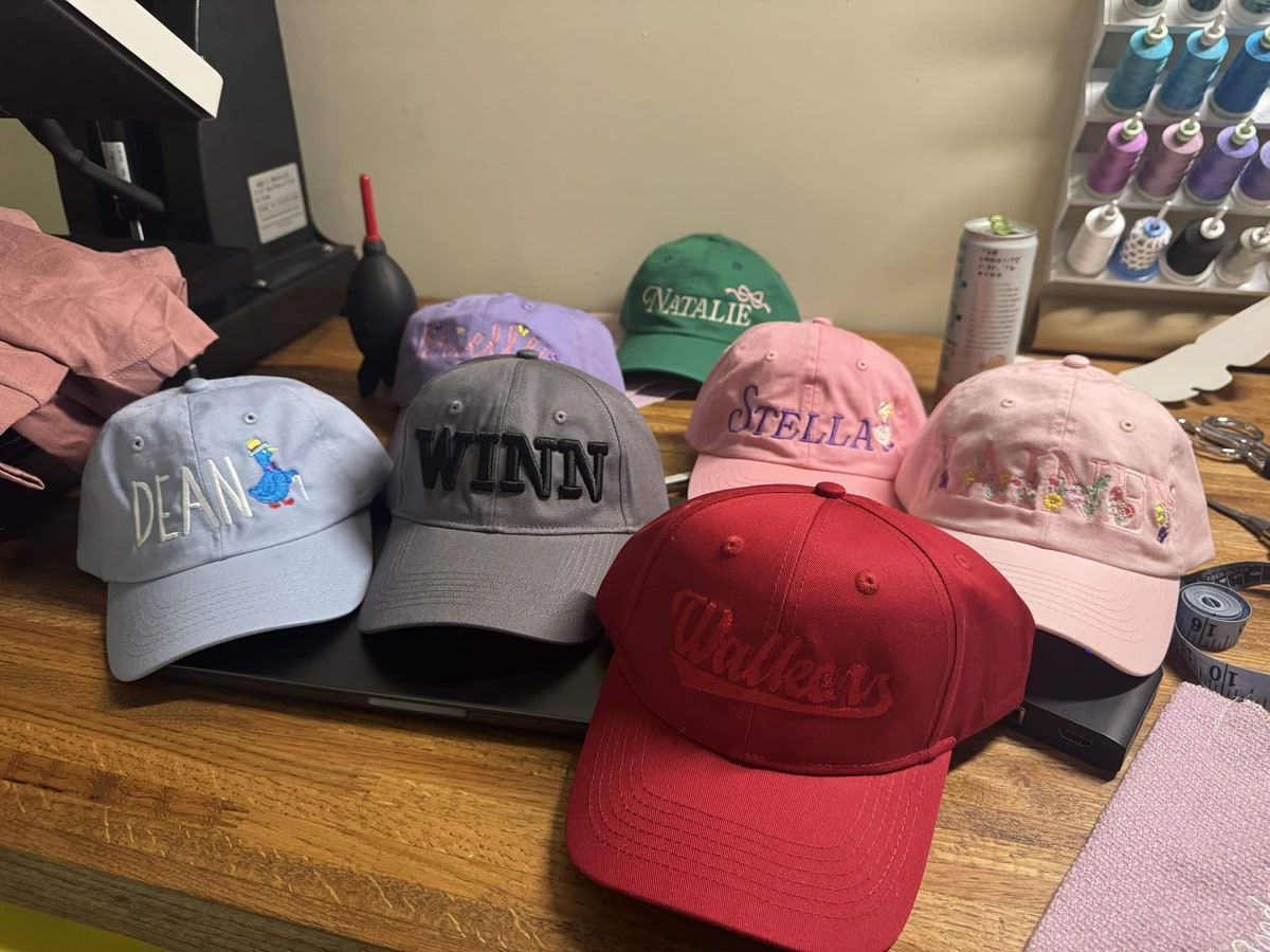 Seven baseball caps embroidered with personalised names, on a workshop table beside a rack of thread cones.