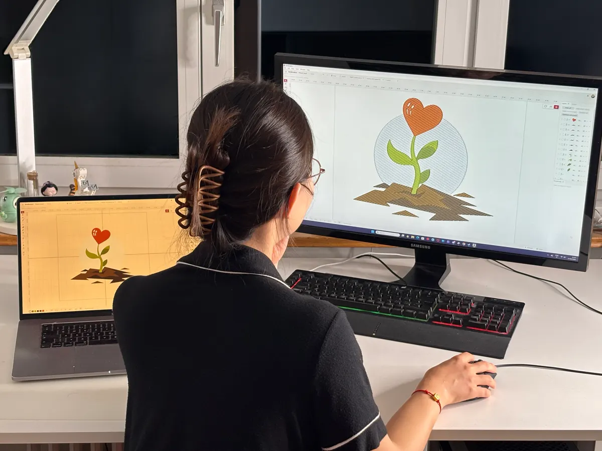 The same person at a desk, the same heart-and-flower design open in Boldbobbin on a wide monitor and on the laptop beside it, one hand on the mouse.