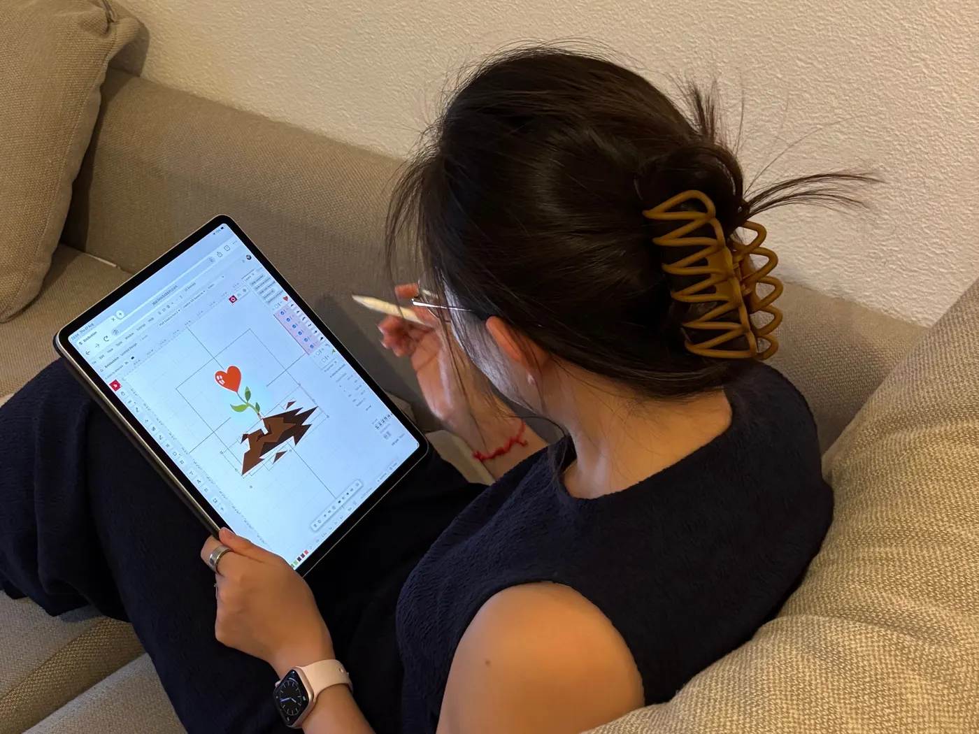 Someone sitting on a sofa with a tablet on their knee, drawing on a heart-and-flower embroidery design in Boldbobbin with a stylus.