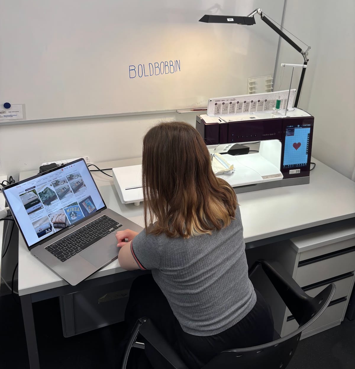 A maker at a desk scrolling a marketplace full of other people's embroidery designs on a laptop, with her own embroidery machine idle beside her.
