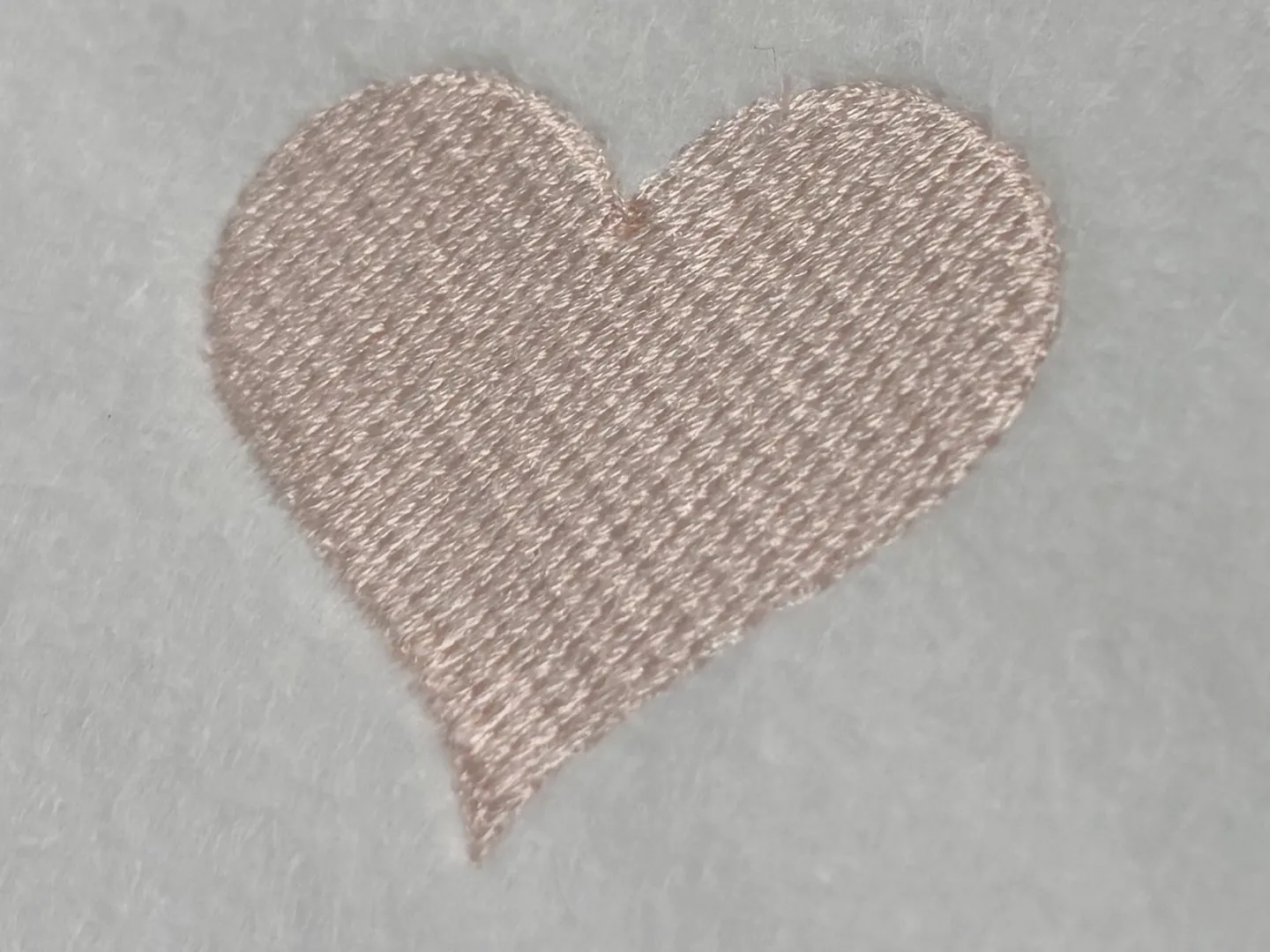 The heart sewn in pale pink thread on white fabric, close enough to read the rows of stitches running across it.