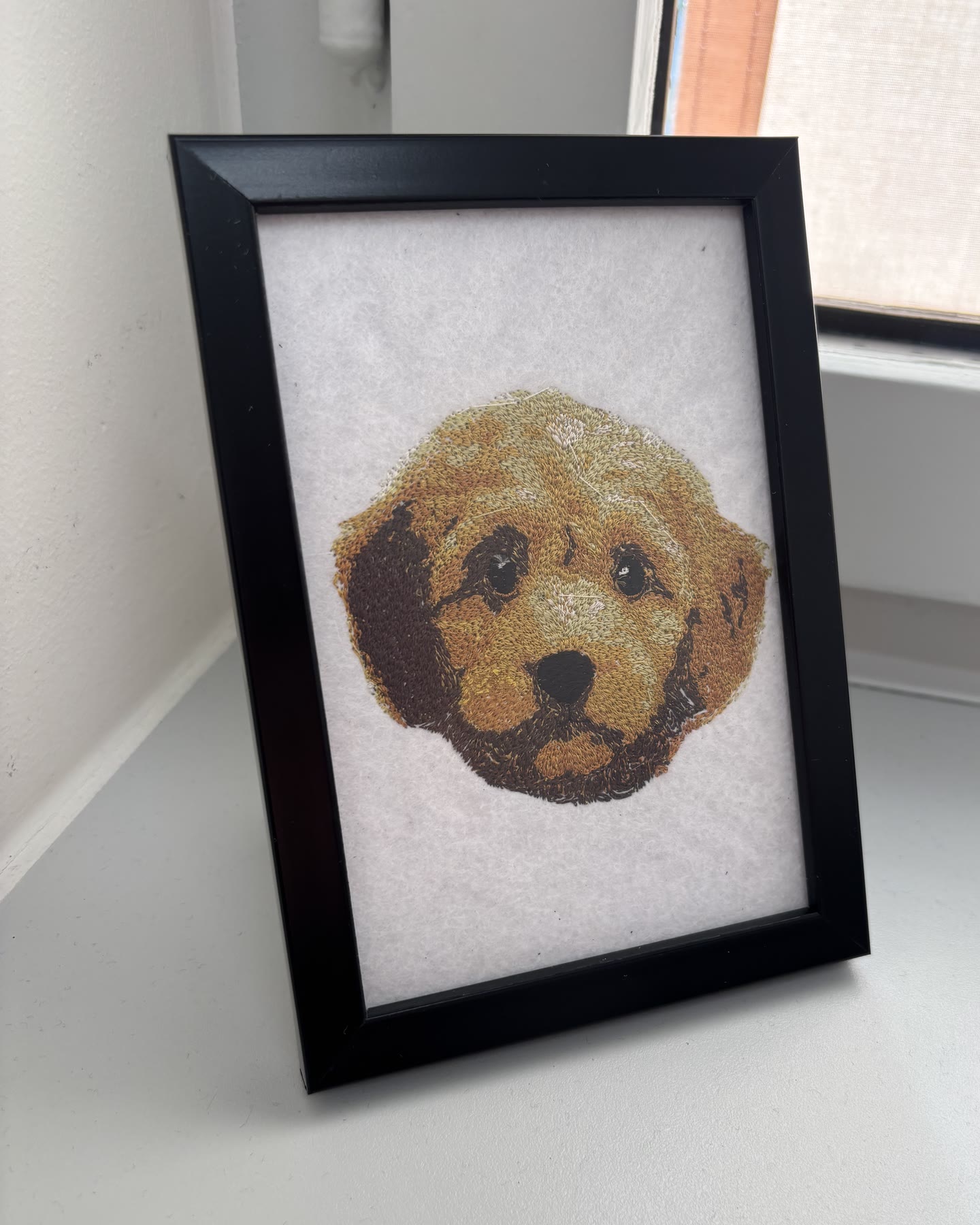 The finished golden puppy embroidery in a black frame, standing on a windowsill.