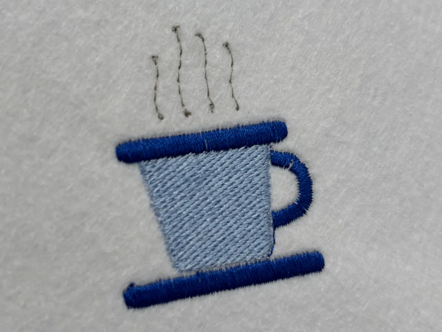The finished coffee cup sewn on white fabric: a pale blue body, a dark blue rim, saucer and handle, and four grey wisps of steam.