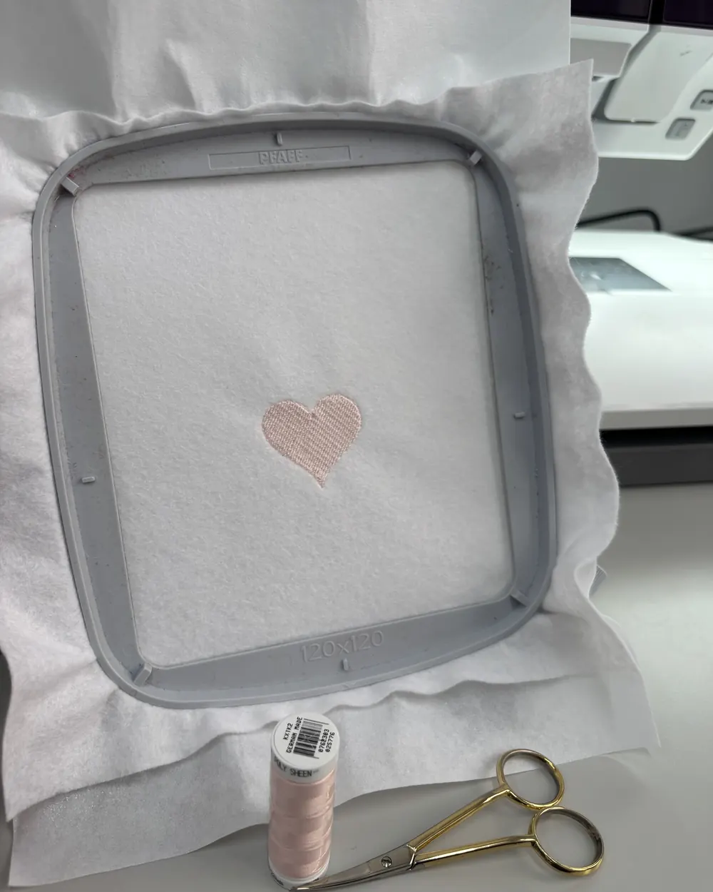 The finished heart stitched in pale pink, still in the hoop, with the spool it was sewn from and a pair of embroidery scissors beside it and the machine behind.