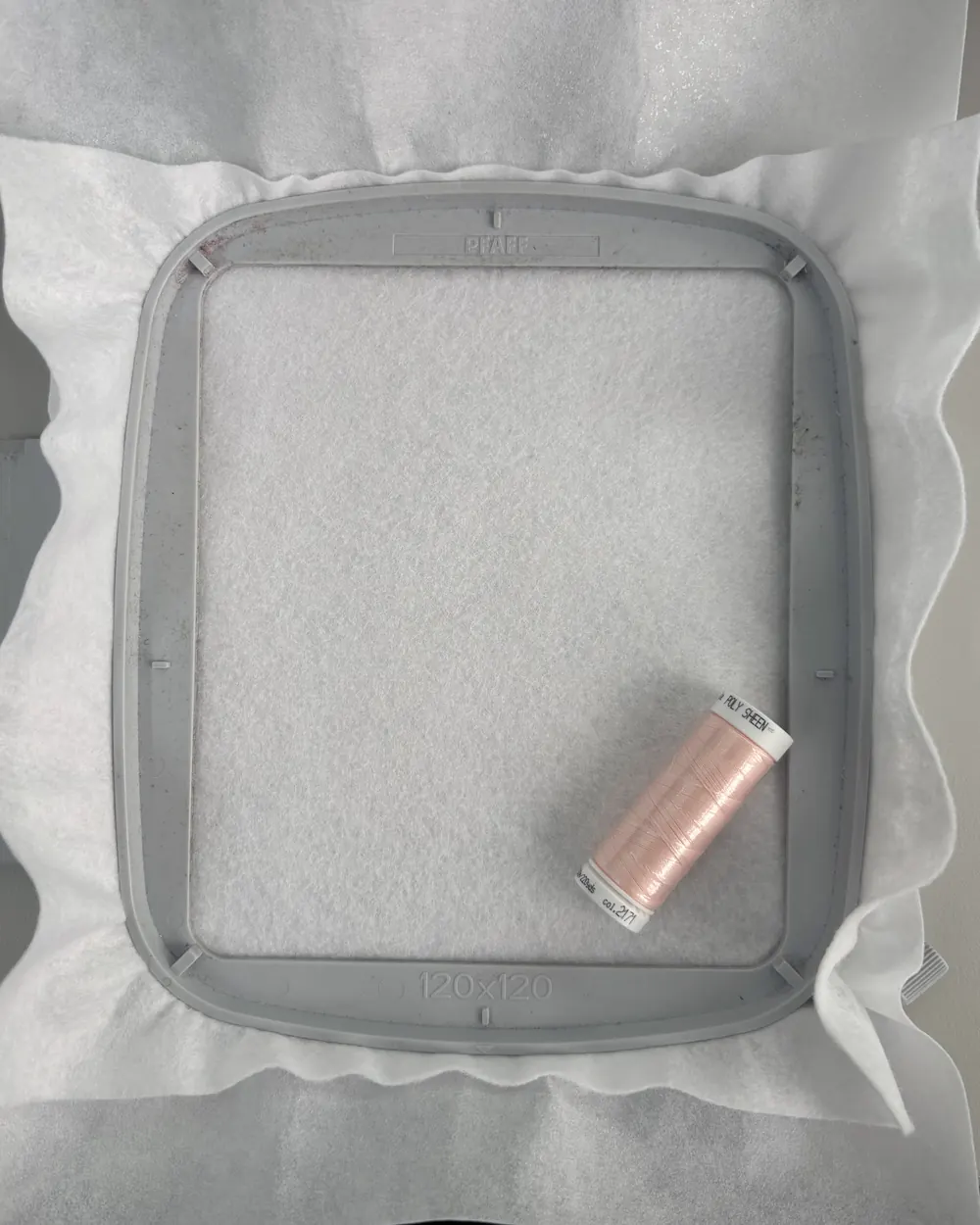 White fabric and stabiliser clamped together in a square Pfaff 120 by 120 hoop, the spare material spreading out past the frame, with a spool of pale pink thread resting on top.