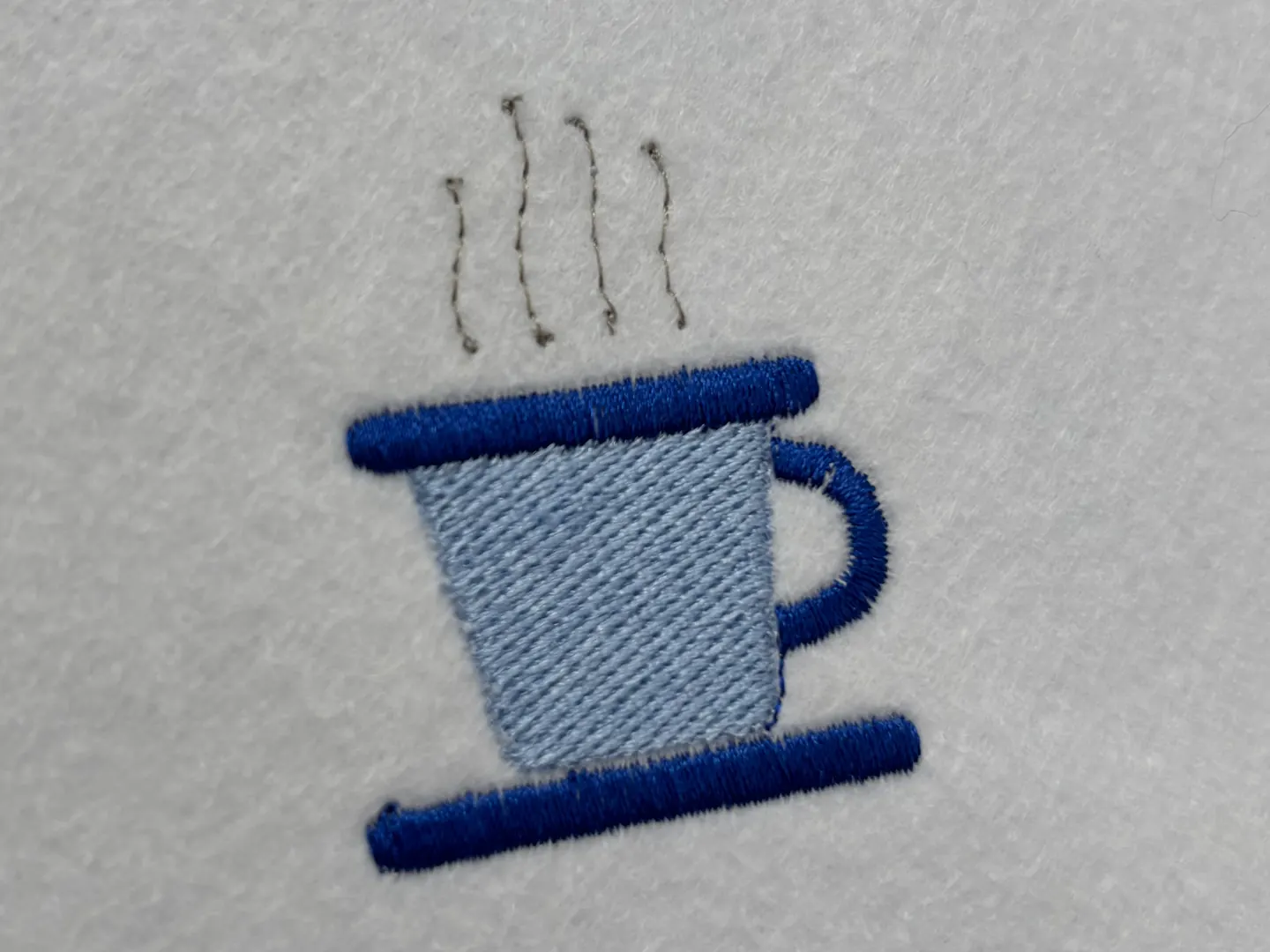 The finished coffee cup in thread: a pale blue body filled with angled rows, dark blue bands for the rim, saucer and handle, and four grey wisps of steam above it.