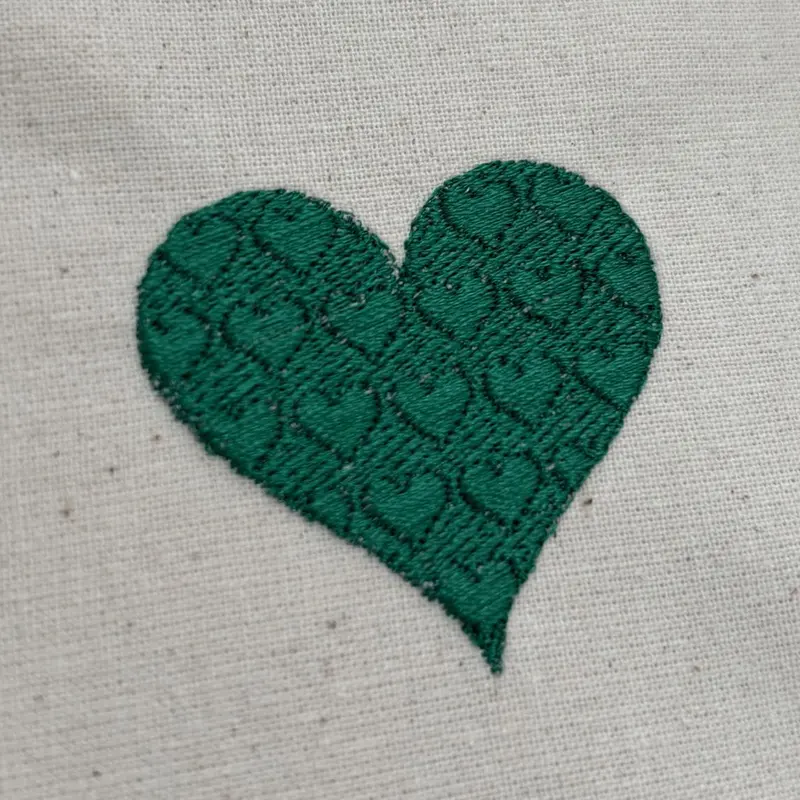 A heart embroidered in green thread on cream fabric, its surface textured with a repeating pattern of small hearts worked into the fill.